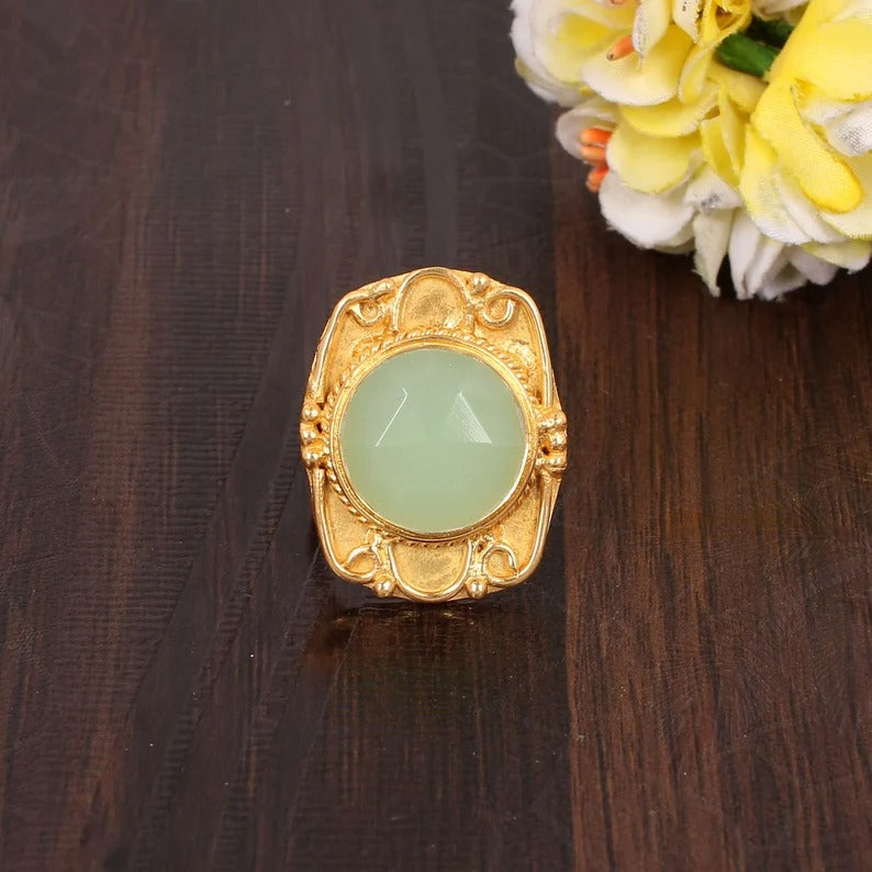 A gold-plated brass ring with a round cut prehnite chalcedony gemstone, placed on a dark surface with a floral arrangement in the background.